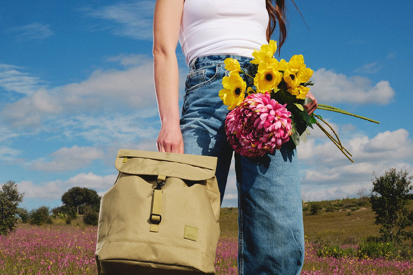 Person holding a beige backpack and flowers in a field with a blue sky.