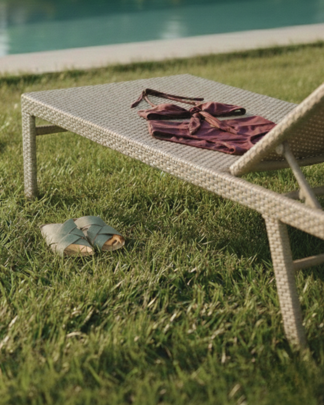 Wicker sun lounger with a bikini and sandals on grass near a pool