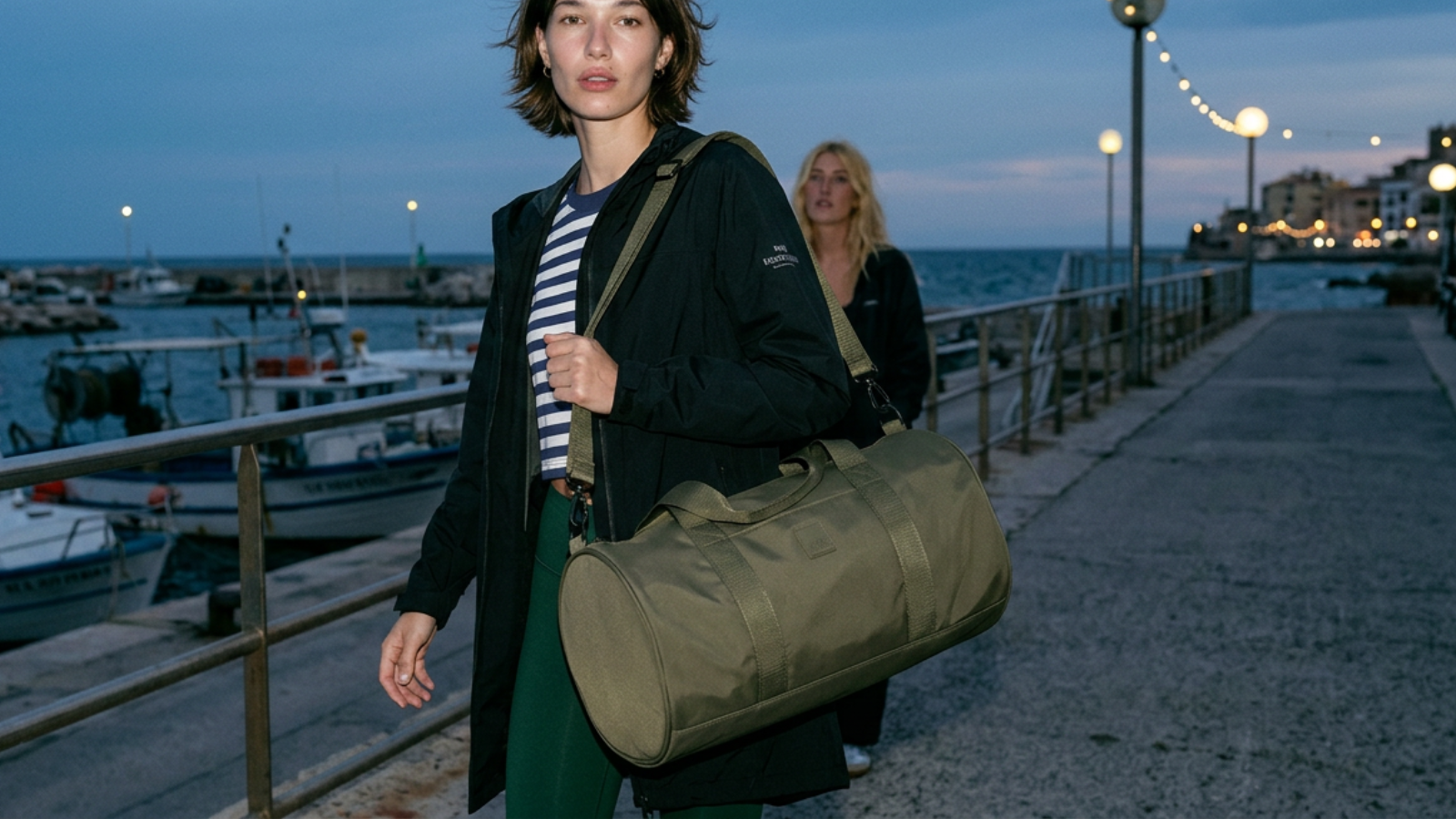 Woman walking along a waterfront promenade with a green duffel bag.