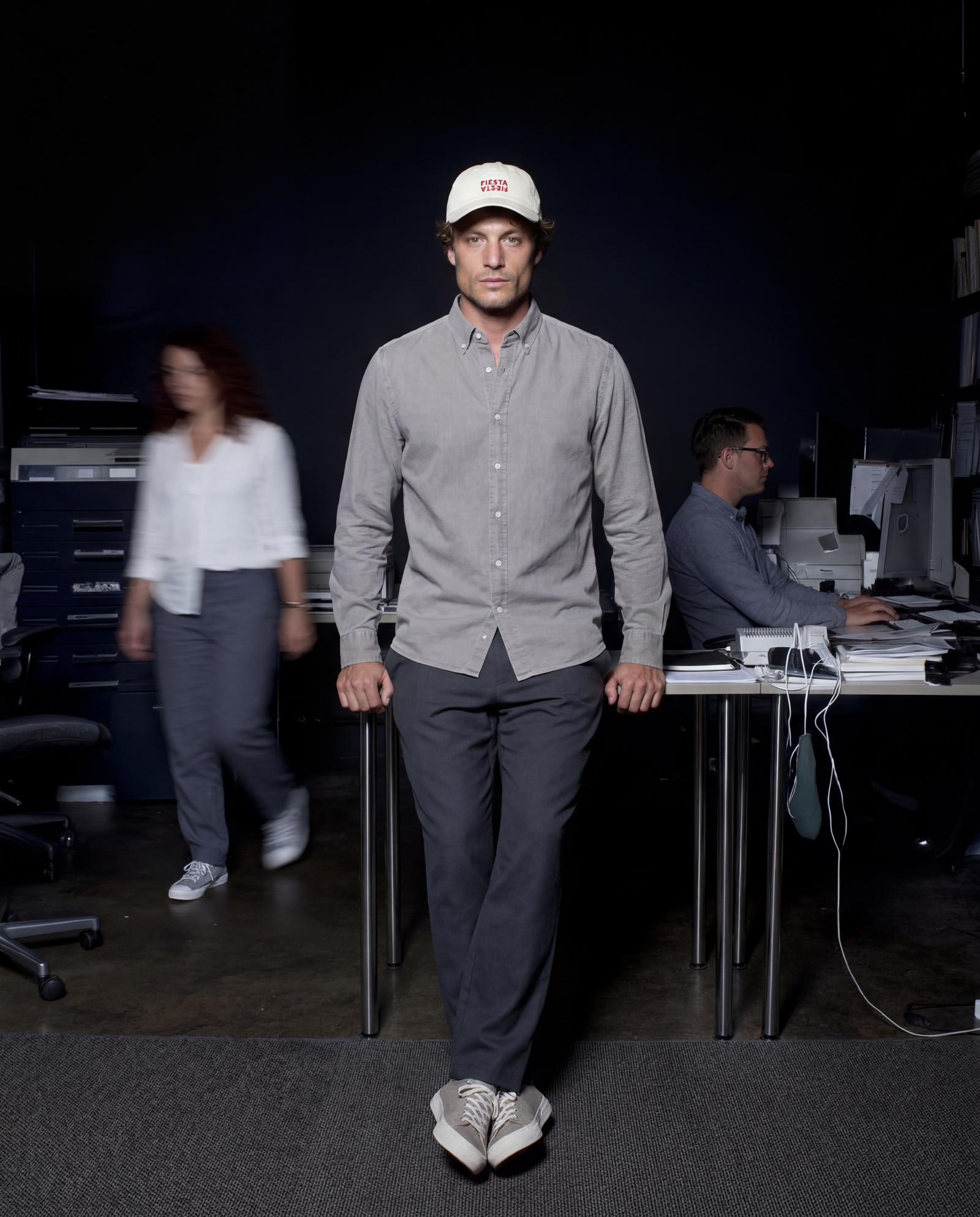 A man walks forward in a dim office space wearing a light gray button-down shirt, dark gray trousers, white sneakers, and a cream baseball cap with a red logo. Two blurred people and office desks are visible in the background.