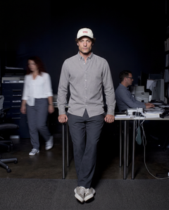 A man walks forward in a dim office space wearing a light gray button-down shirt, dark gray trousers, white sneakers, and a cream baseball cap with a red logo. Two blurred people and office desks are visible in the background.