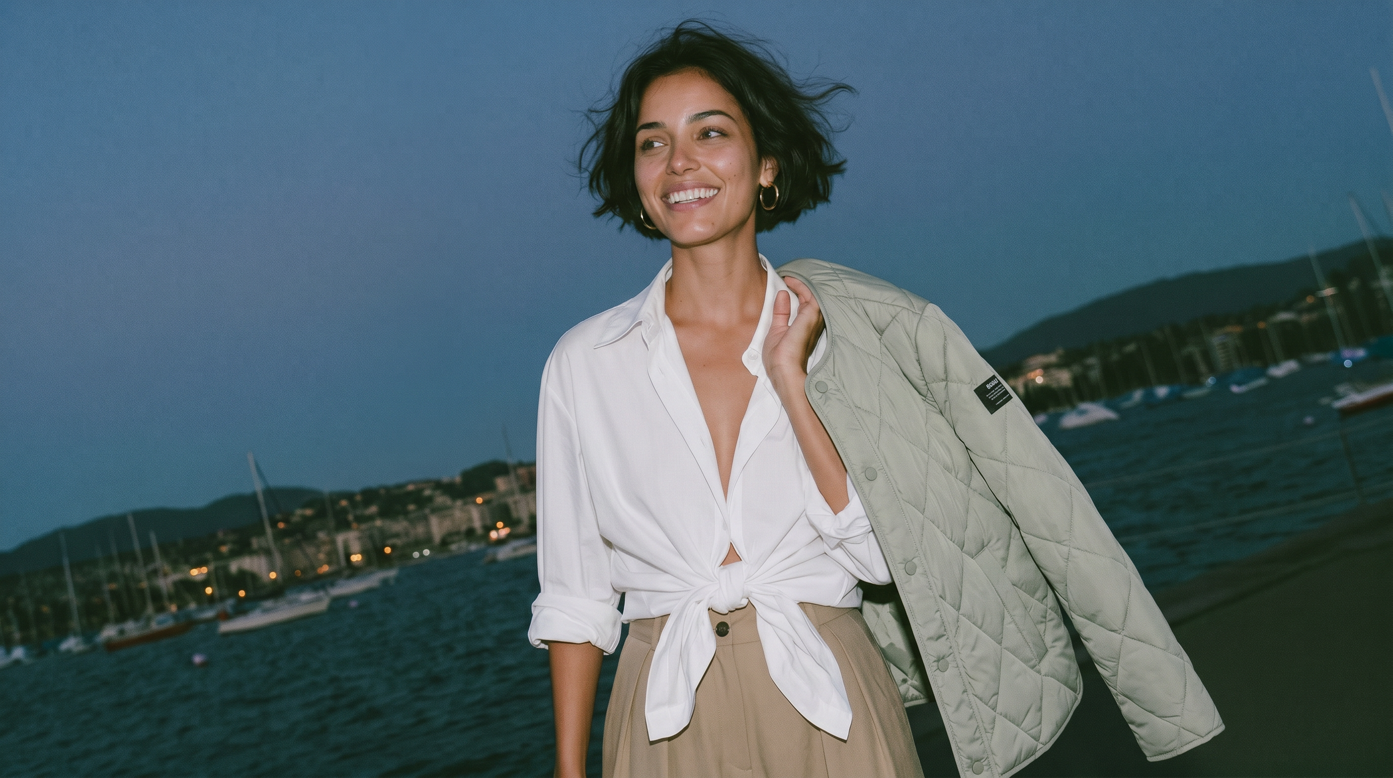 A woman standing outdoors by the water at dusk, wearing a white tied button-up shirt, beige skirt, and holding a light beige quilted jacket over her shoulder.