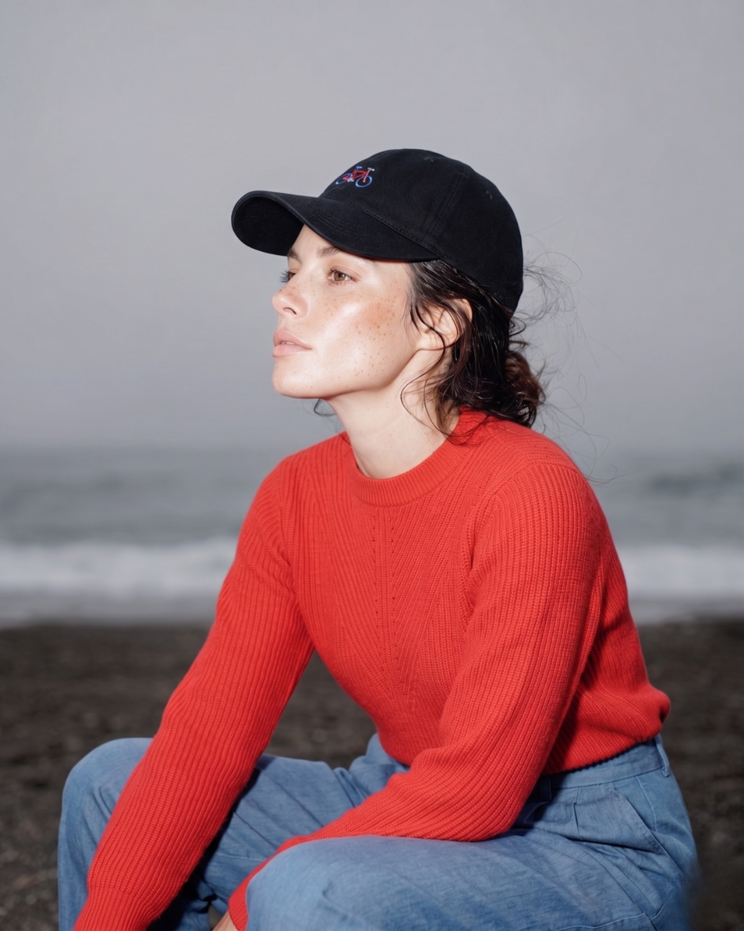 Person wearing a red sweater and black cap sitting on a beach.
