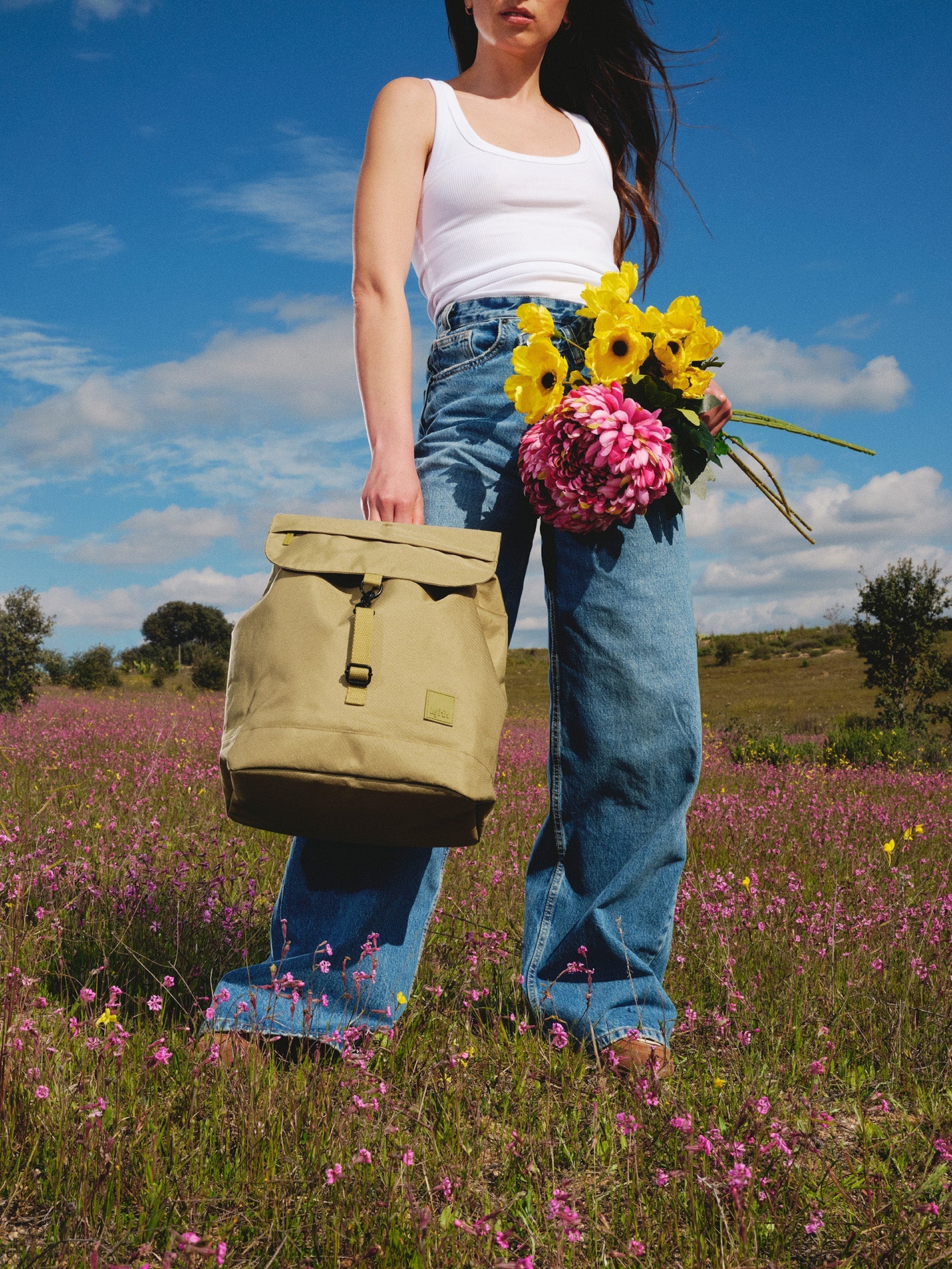 Person holding a beige backpack and flowers in a field with a blue sky.